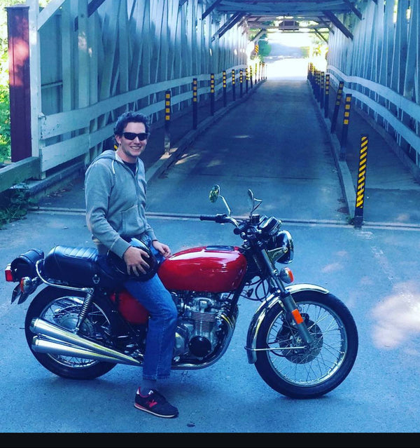 A man wearing sunglasses sits on a red classic motorcycle on the left side of a bridge, with the motorcycle positioned in front of him. The bridge has a white metal truss structure and yellow-and-black striped safety posts along the sides.