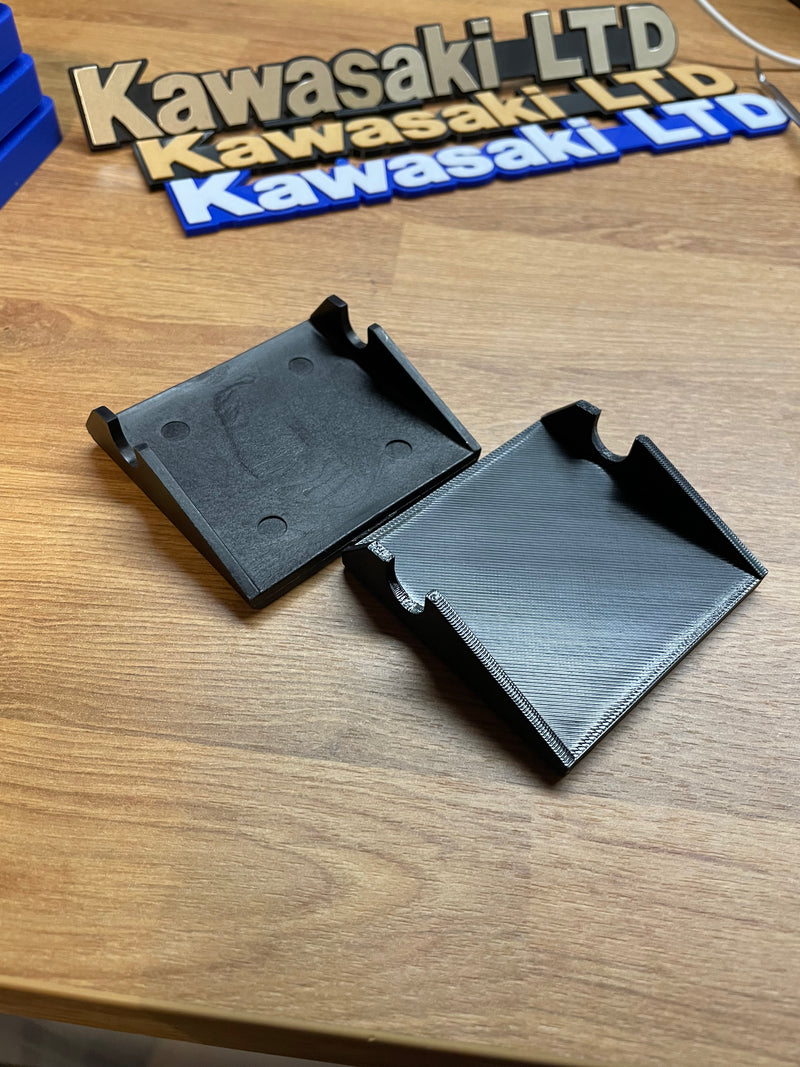 Two black 3D-printed plastic mounting brackets or covers displayed on a wooden tabletop, with a Kawasaki LTD logo sign visible in the background.